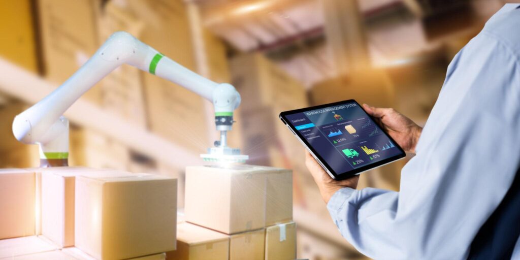 Bras robotisé manipulant un colis dans un entrepôt automatisé tandis qu’un opérateur supervise les stocks sur une tablette numérique.